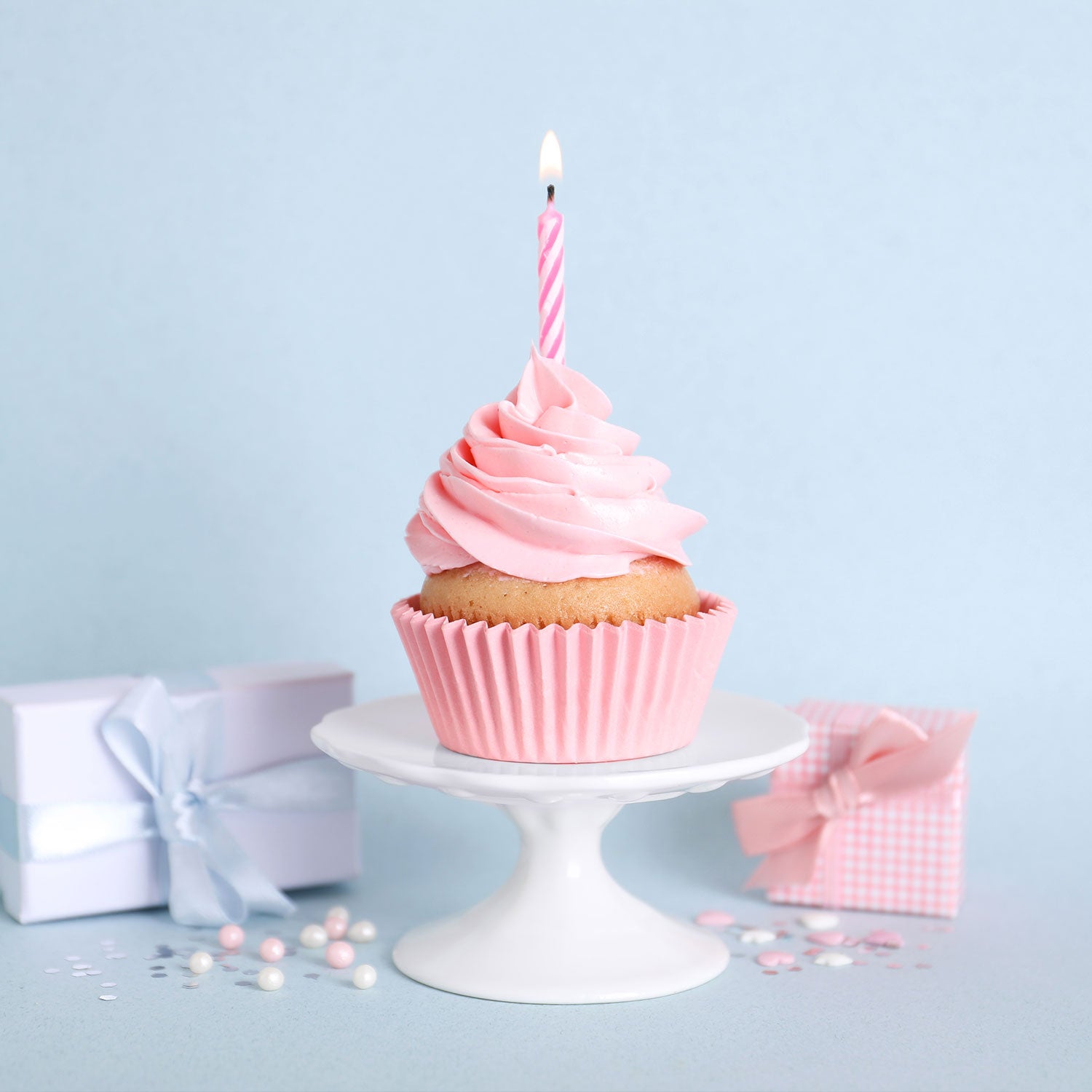 Pink cupcake with a lit candle on a white cake stand against a light blue background