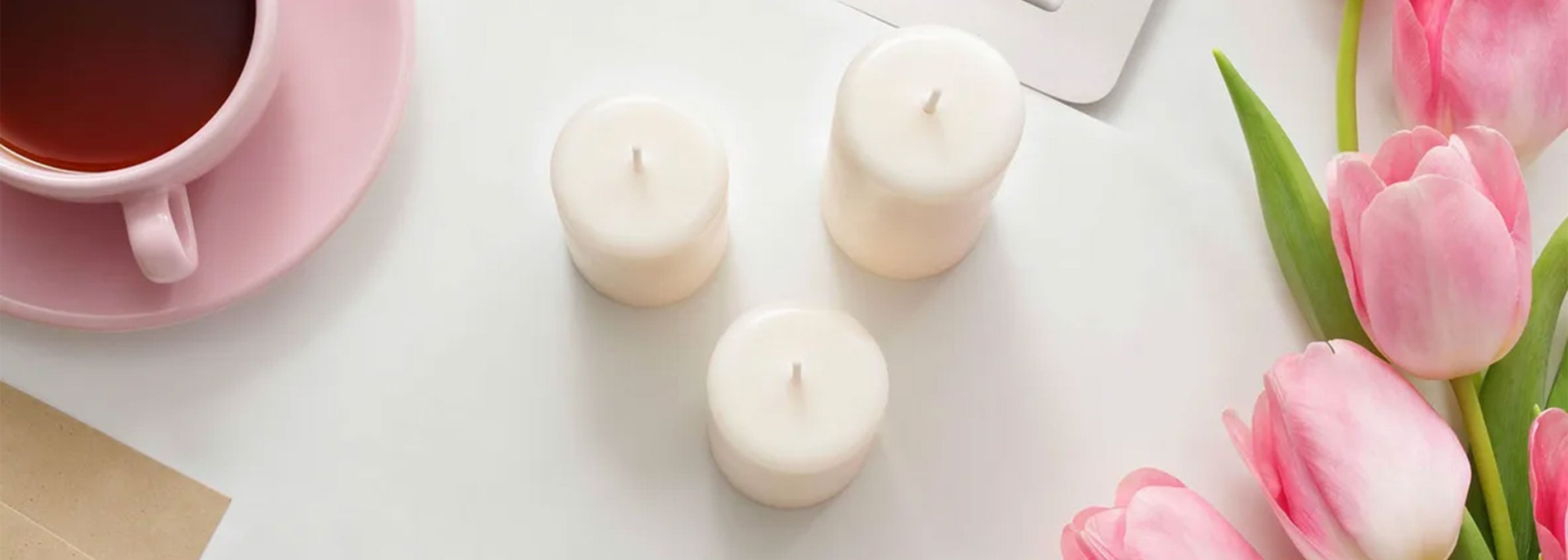Candle holder with three white candles, a pink cup of tea, and pink tulips on a white surface.