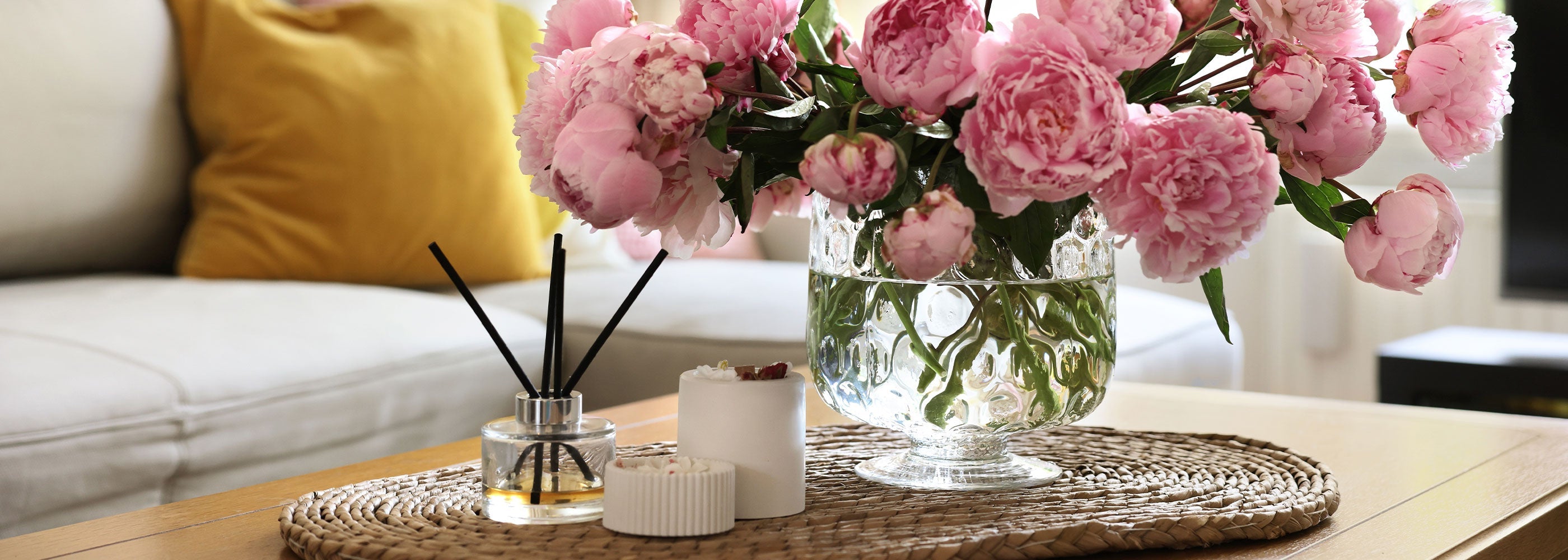 Pink flowers in a vase on a coffee table with a diffuser and candles, next to a yellow pillow on a couch.